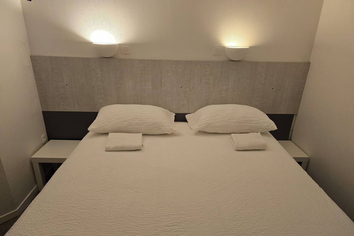 A neatly made bed with two pillows and two folded towels, flanked by two bedside tables with wall-mounted lamps.