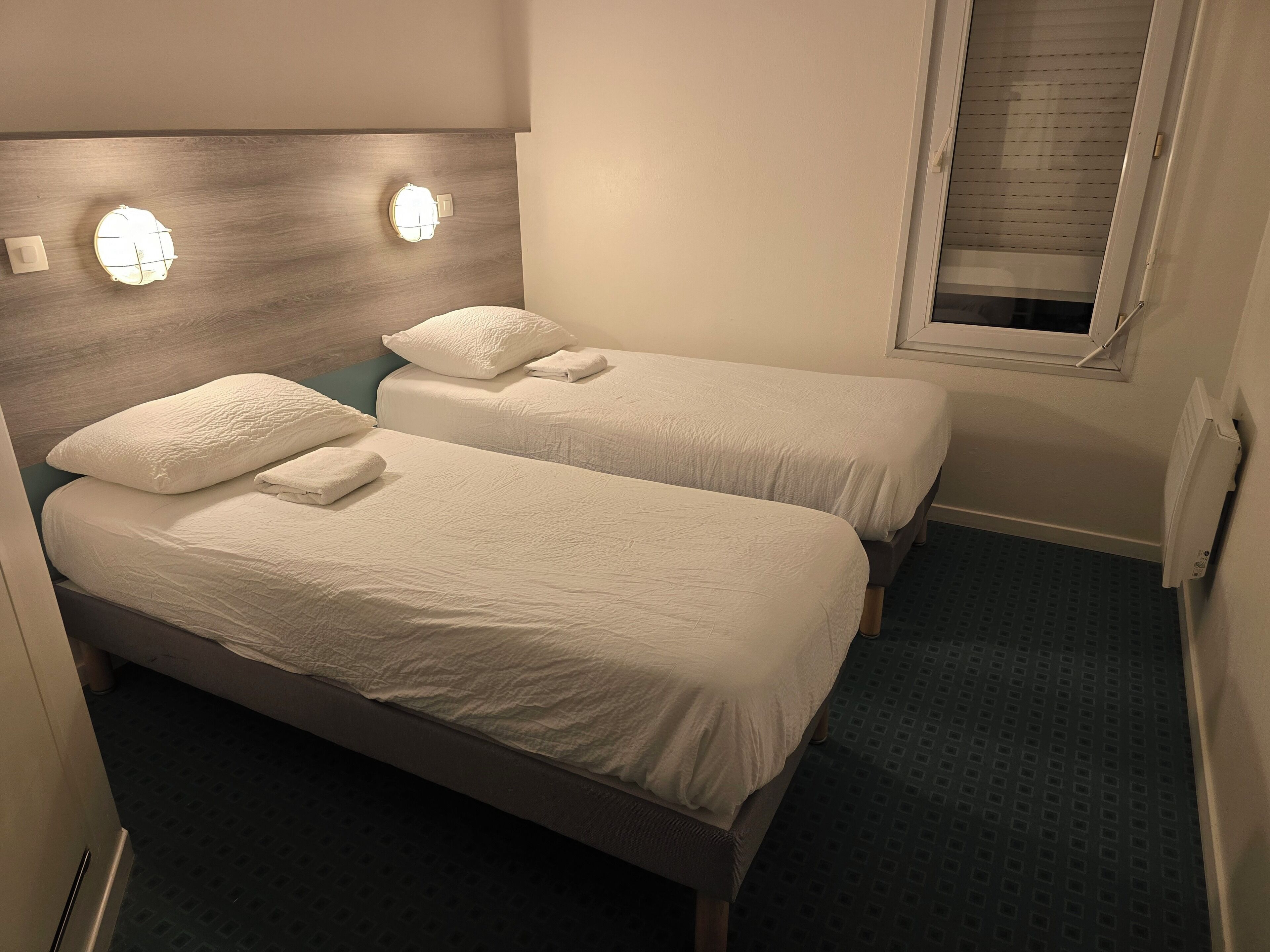 Ein Hotelzimmer mit zwei Betten, jeweils mit weißen Leinen und Kissen, einem Holz-Kopfschild und einem Fenster mit Jalousien.