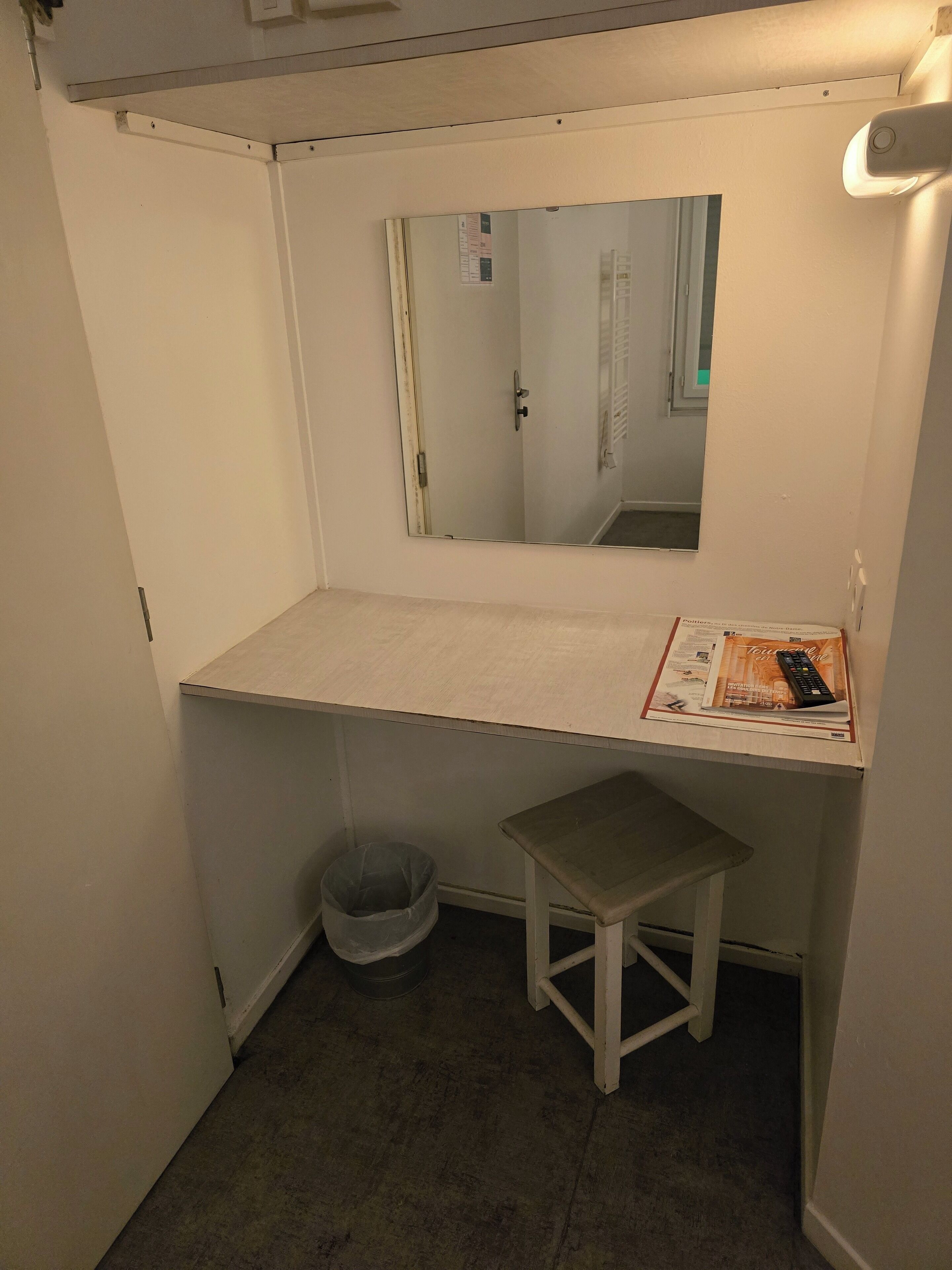 A small, narrow room with a white desk, a mirror, and a small stool.