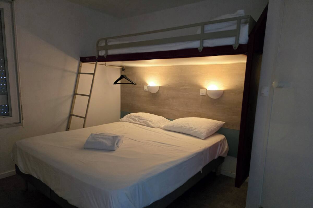 A single bed with a bunk above, a ladder, two wall-mounted lights, and a window with blinds.