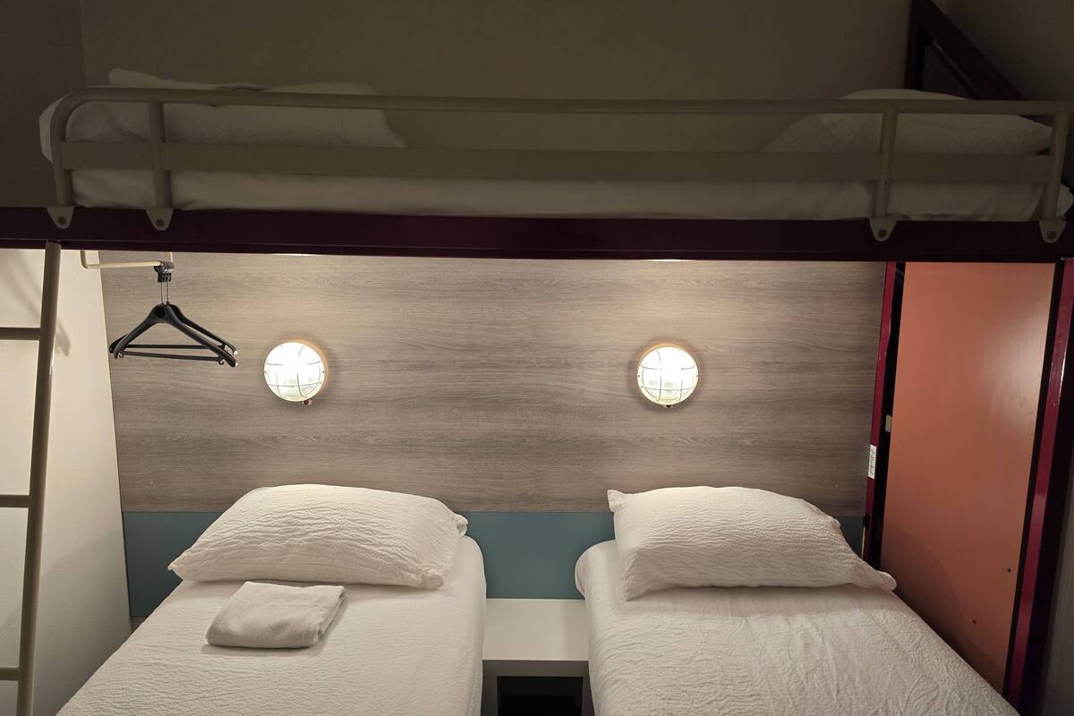 A double room with two beds, each with a pillow and a folded towel. There are two wall-mounted lights and a wooden headboard.