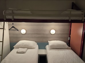 A double room with two beds, each with a pillow and a folded towel. There are two wall-mounted lights and a wooden headboard.