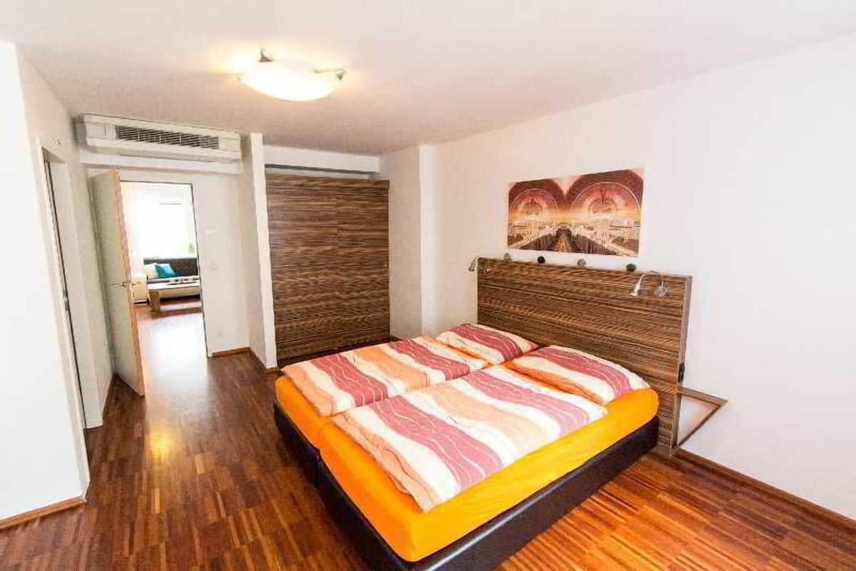 Quarto com cabeceira de madeira, cama com roupa de cama listrada, pintura na parede e corredor que dá para outro quarto.