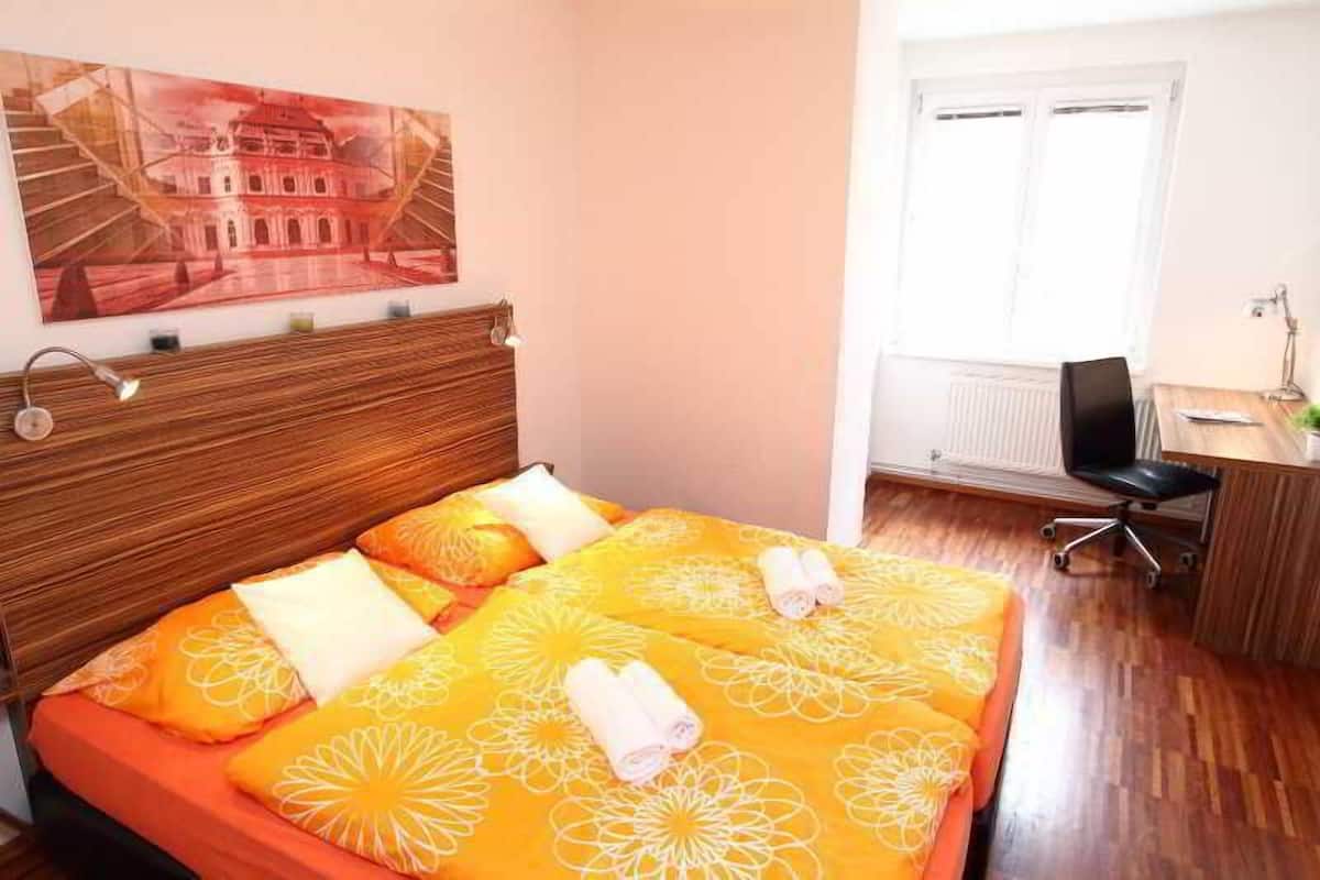 Quarto de hotel com cama, escrivaninha, cadeira, janela e um quadro na parede.