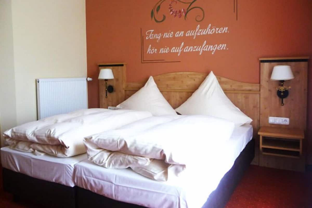 Ein Hotelzimmer mit einem Doppelbett, zwei Nachttischen mit Lampen und einer Wand mit einer Dekoration und Text auf Deutsch.