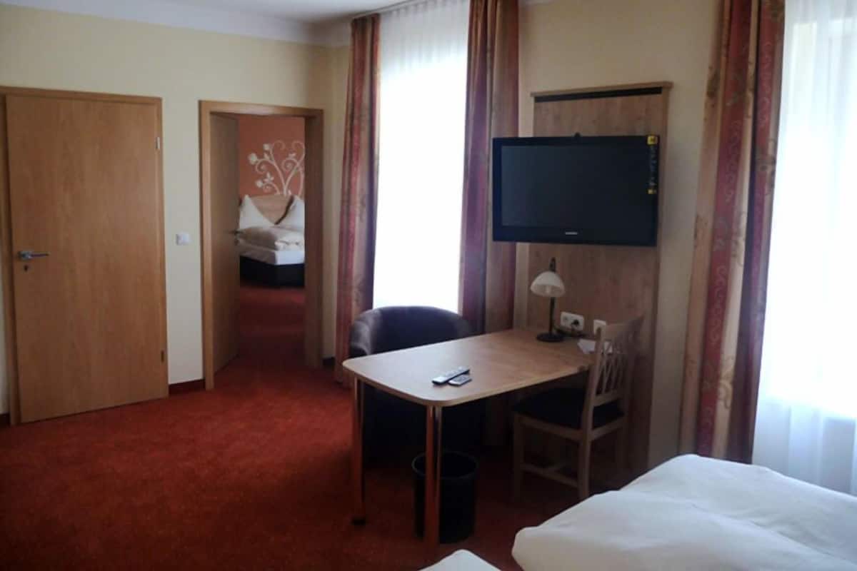 Ein Hotelzimmer mit einem Bett, einem Schreibtisch mit Stuhl, einem Fernseher und einer Tür zu einem anderen Raum.