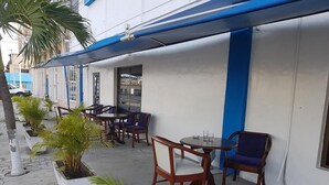 Uma área externa com mesas e cadeiras, plantas em vasos e um edifício com paredes externas azuis e brancas.