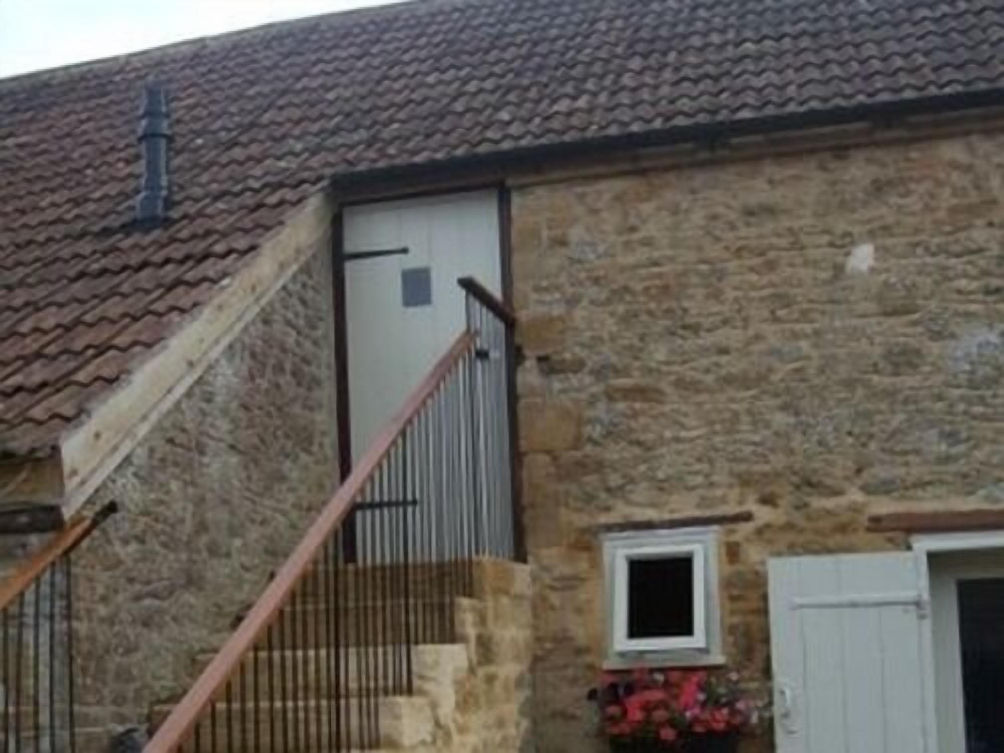 A stone building with a tiled roof, a staircase with metal railings, a door, and a window with flowers.