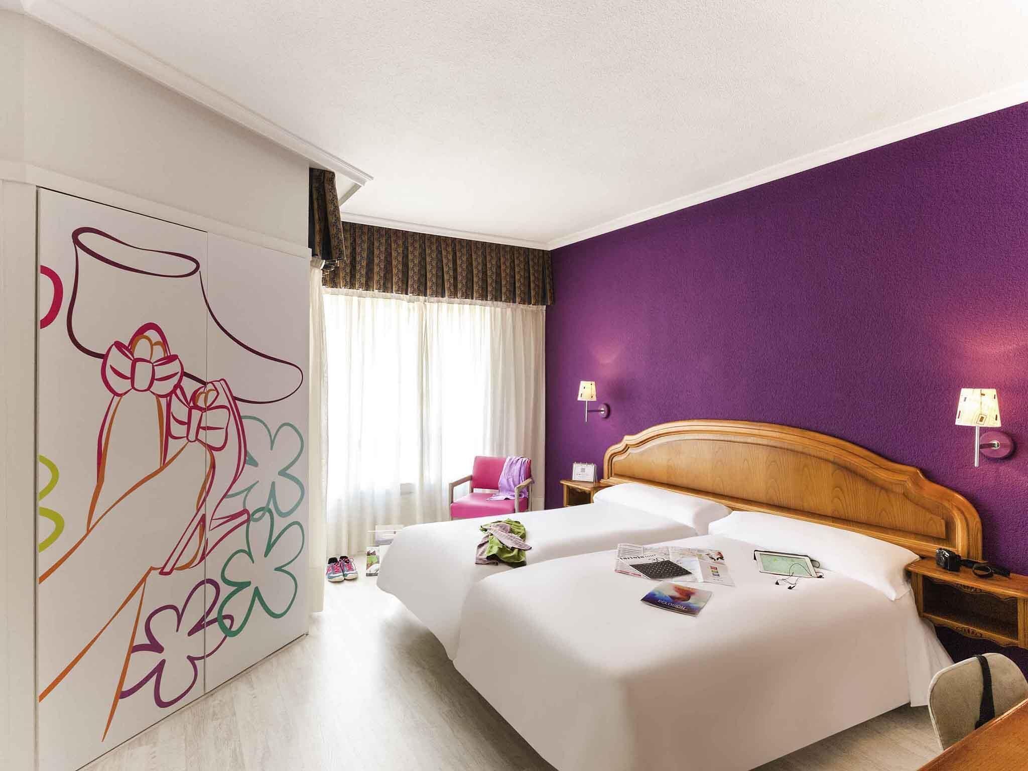 Habitación de hotel con una cama grande, cabecera de madera, pared de acento morada y una ventana con cortinas. Se observa un cuadro abstracto y colorido en la pared.