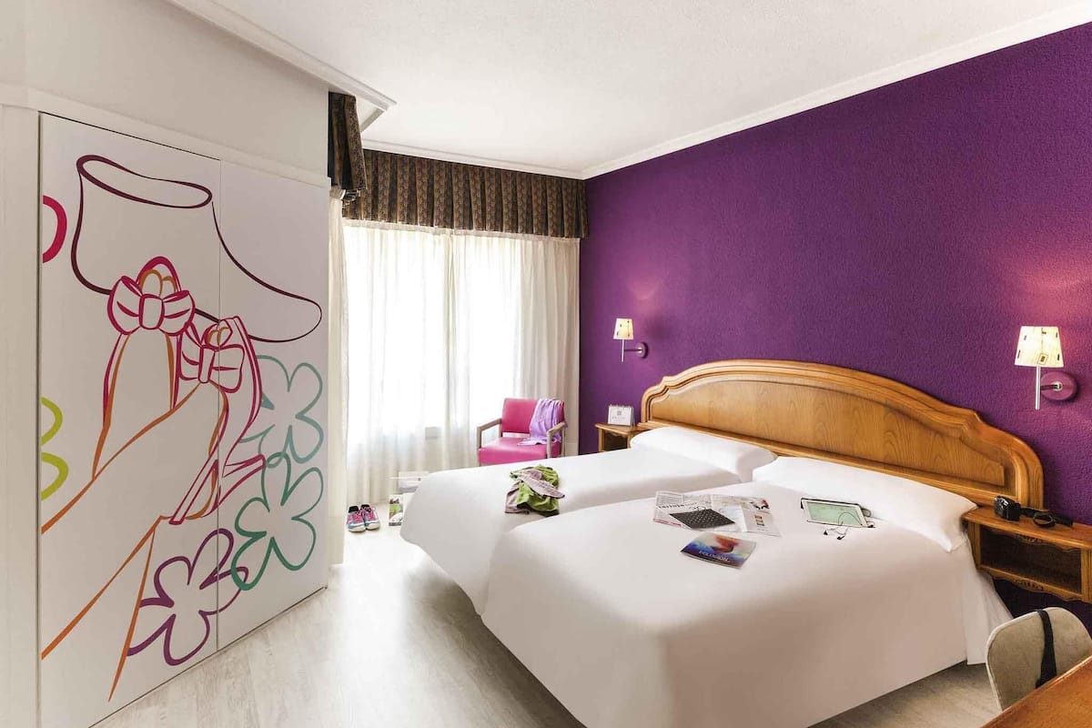 Habitación de hotel con una cama grande, cabecera de madera, pared de acento morada y una ventana con cortinas. Se observa un cuadro abstracto y colorido en la pared.