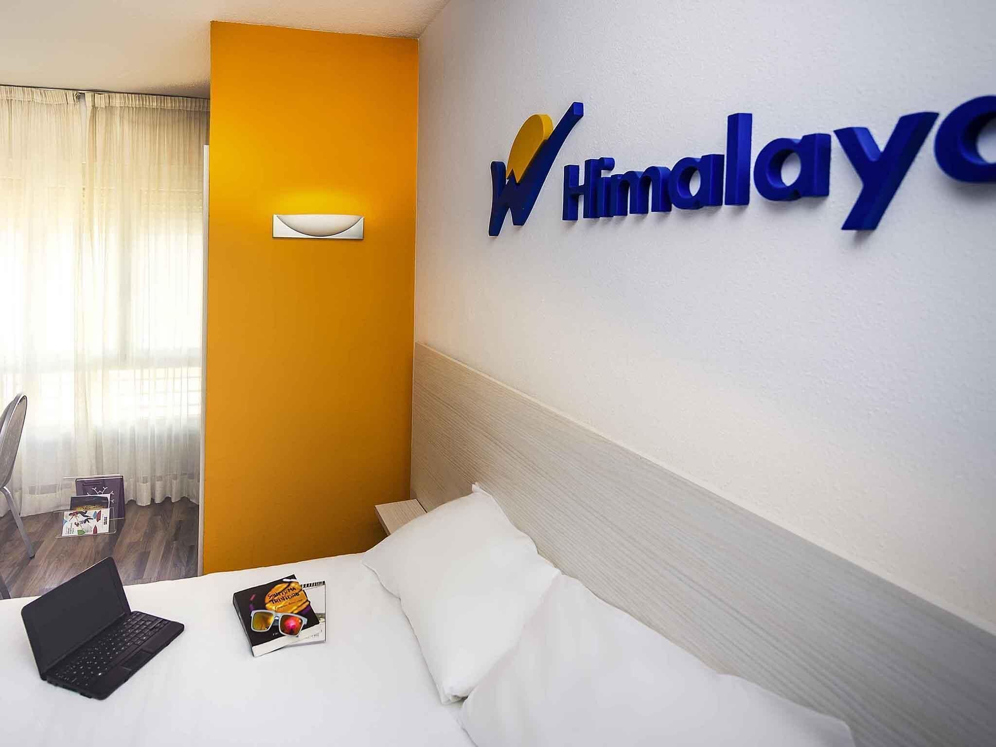 Habitación de hotel con una cama, una computadora portátil, gafas de sol y una revista sobre la cama. Hay un cartel en la pared con el nombre del hotel.