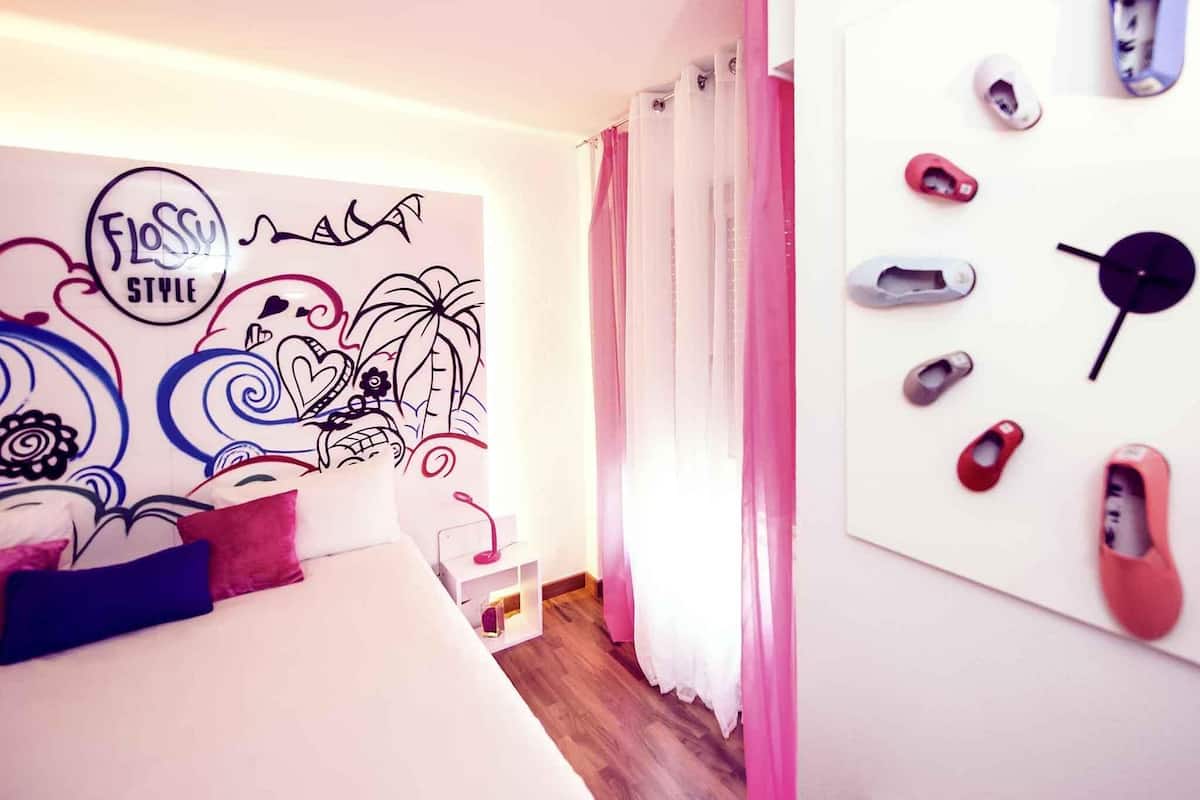 A bedroom with a bed, a wall with a floral and abstract design, a nightstand, and a wall-mounted shoe rack with multiple pairs of shoes.