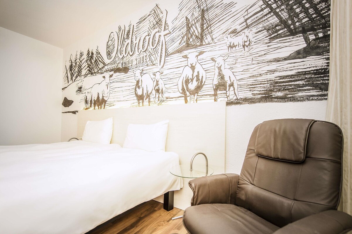 A hotel room with a bed, a brown leather chair, and a wall mural depicting a pastoral scene with sheep and a ship.