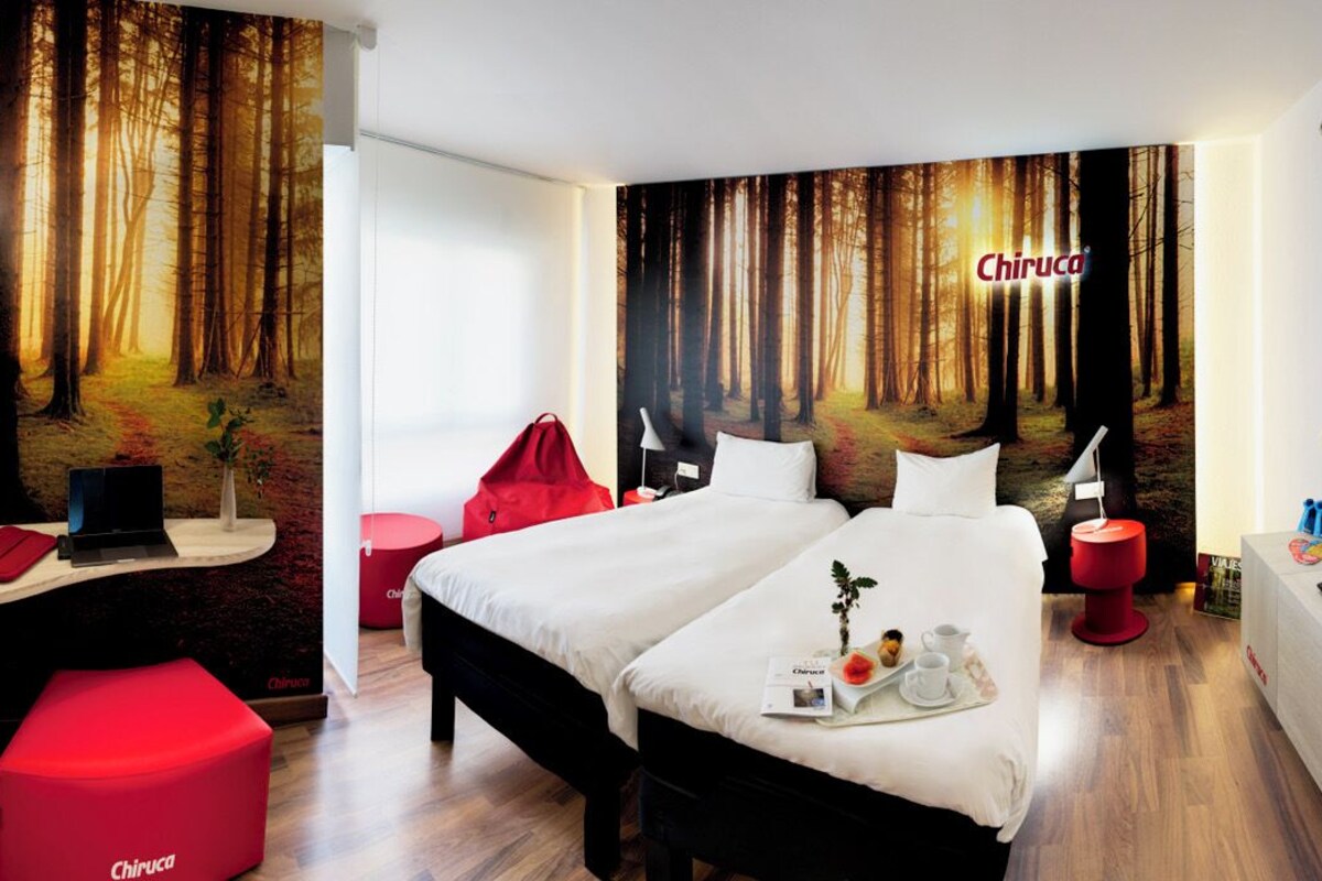 A modern hotel room with a large bed, a desk with a computer, a TV, and a red ottoman.