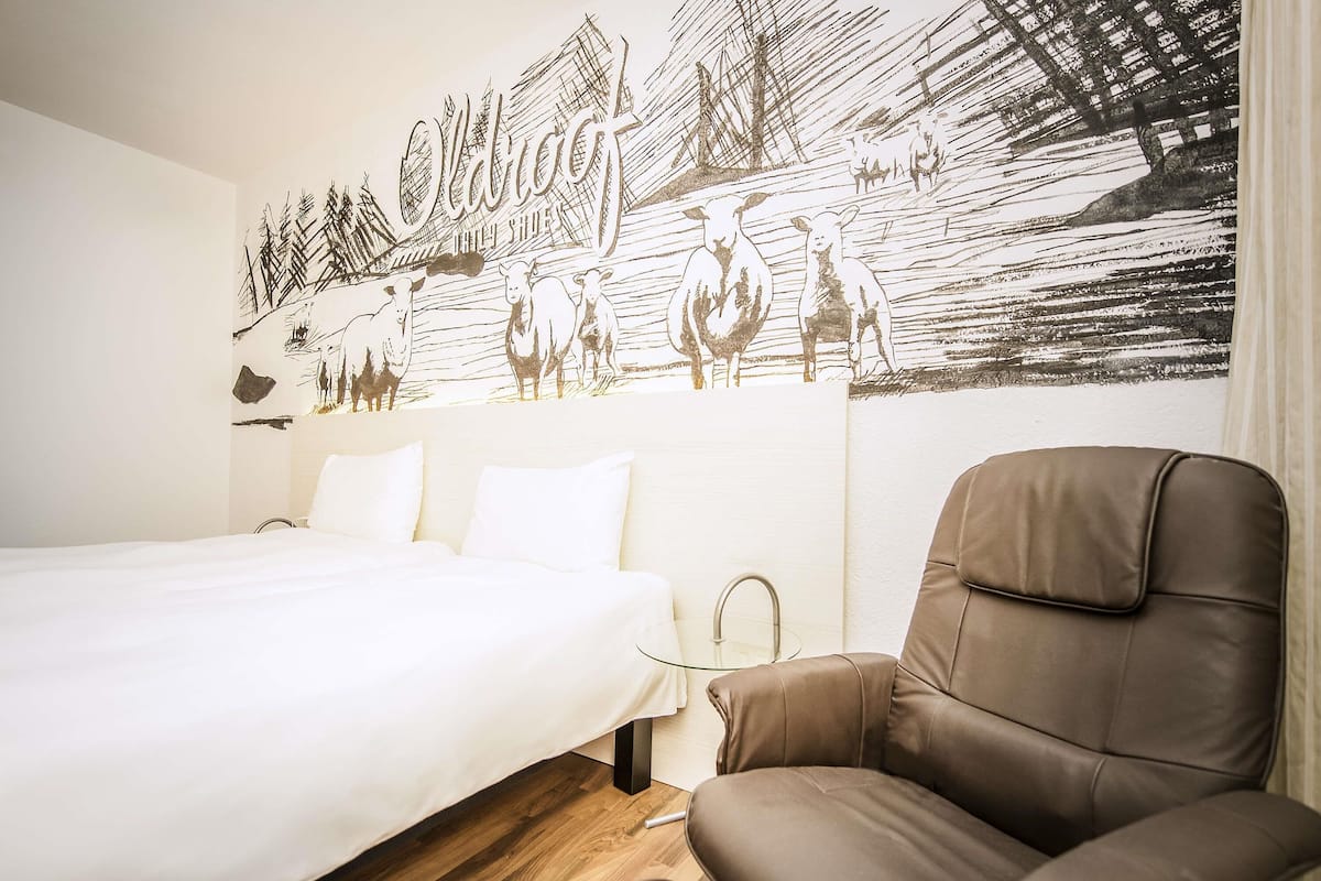 A hotel room with a bed, a brown leather chair, and a wall mural depicting a pastoral scene with sheep and a ship.