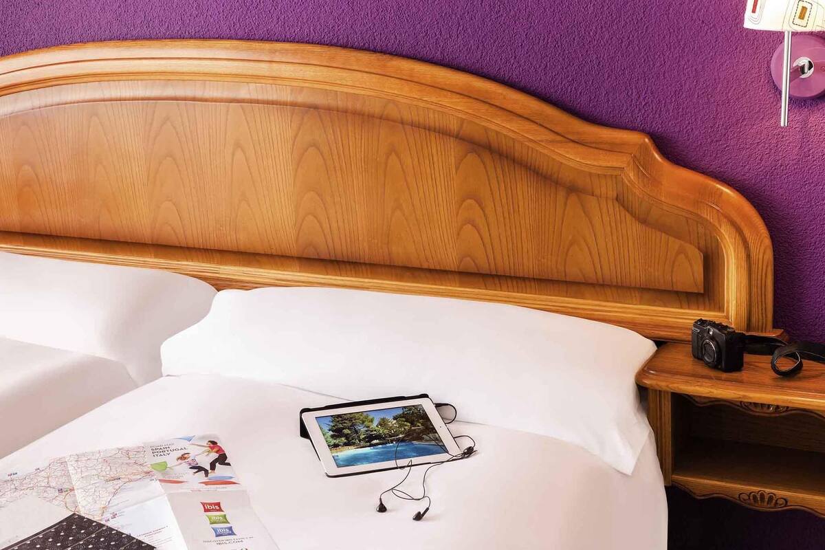 A neatly made bed with a wooden headboard, a white pillow, and a tablet displaying a scenic photo. A camera is placed on the bedside table.