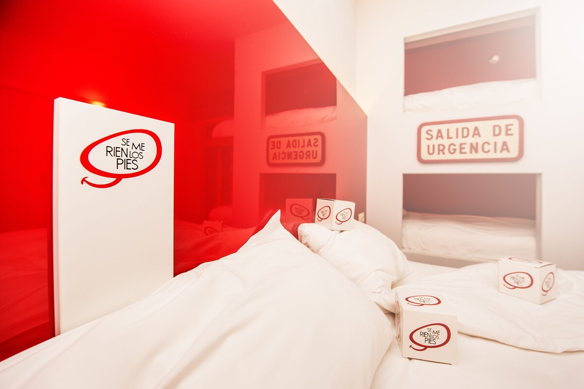 A hotel room with a bed, pillows, and two boxes with a logo. There is a sign on the wall that says "SALIDA DE URGENCIA".