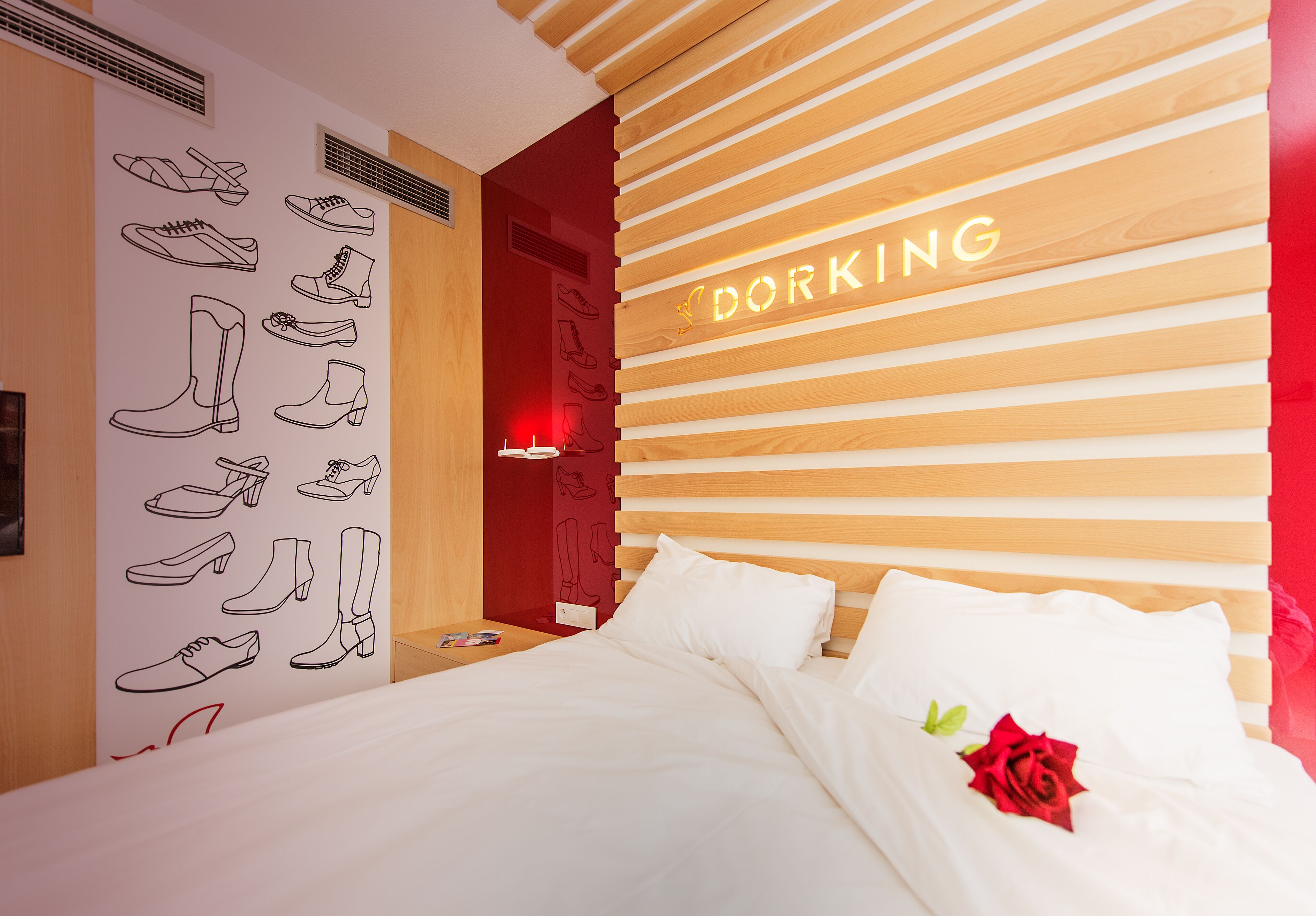 Habitación de hotel con una cama, una pared con ilustraciones de zapatos y un letrero de neón que dice "DORKING".
