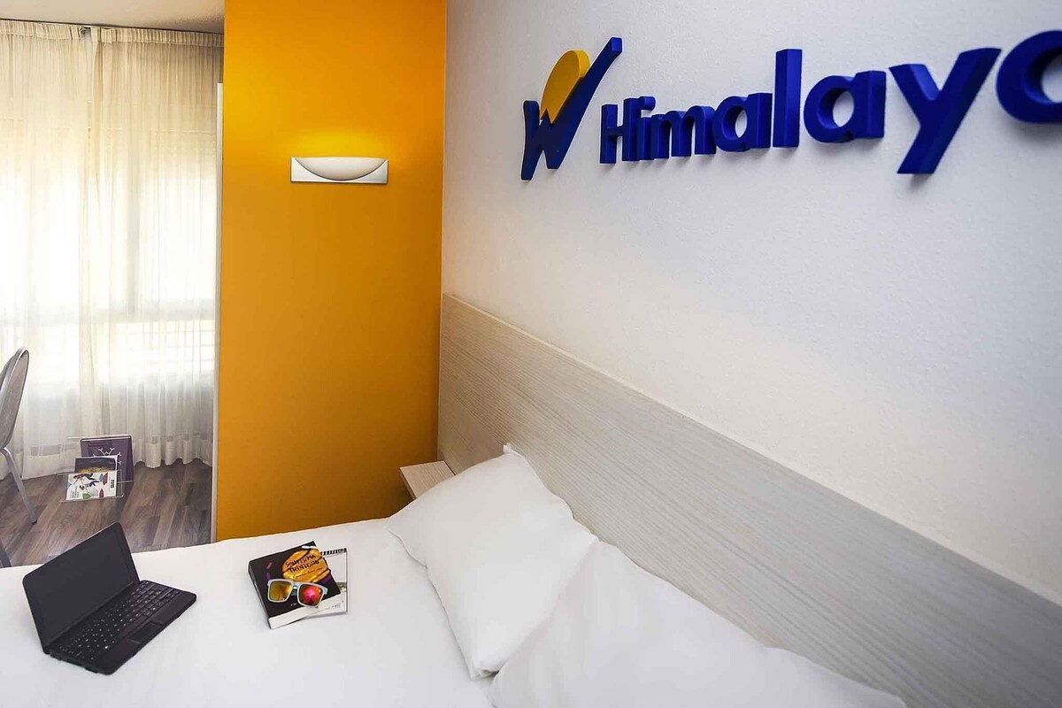 Habitación de hotel con una cama, una computadora portátil, gafas de sol y una revista sobre la cama. Hay un cartel en la pared con el nombre del hotel.