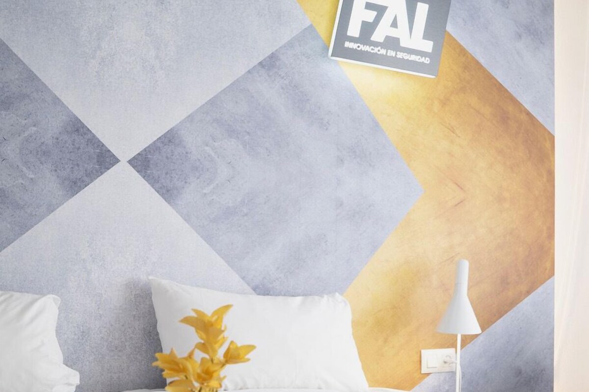 Habitación de hotel con una pared de estampado geométrico, cama con ropa blanca, bandeja con artículos de desayuno y un florero con flores amarillas.