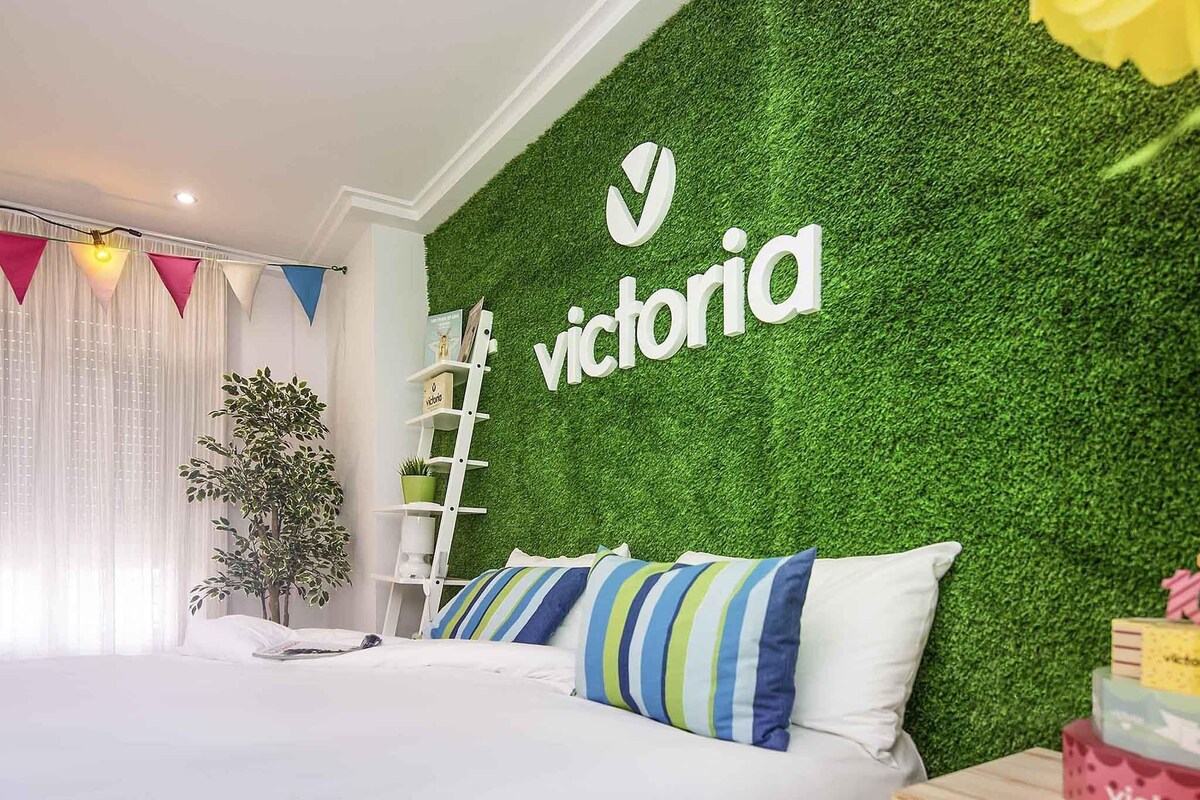 A hotel room with a green wall, a bed with striped pillows, a ladder, and a plant.