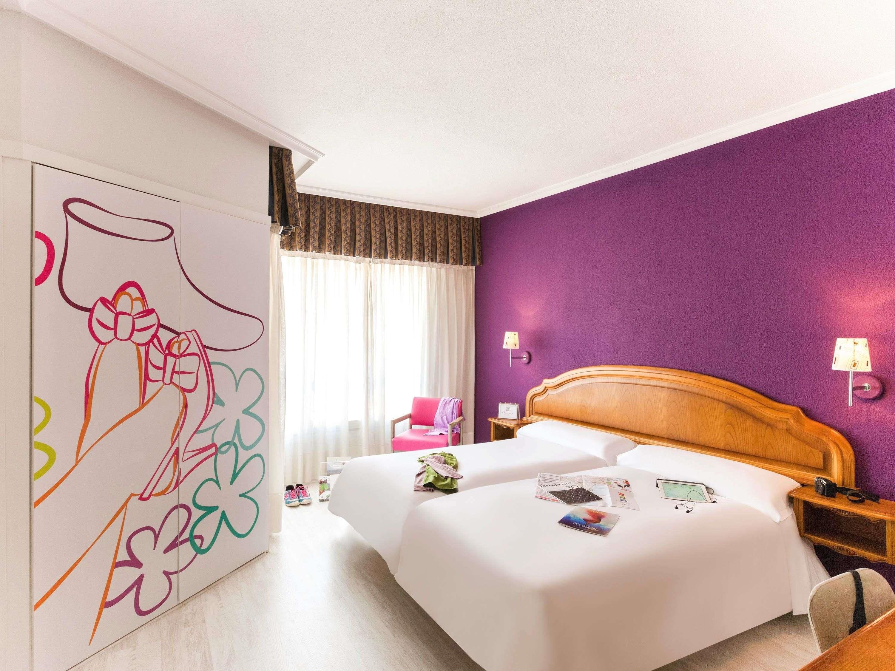 Habitación de hotel con una cama grande, cabecera de madera, pared morada y una obra de arte abstracta colorida en la pared.