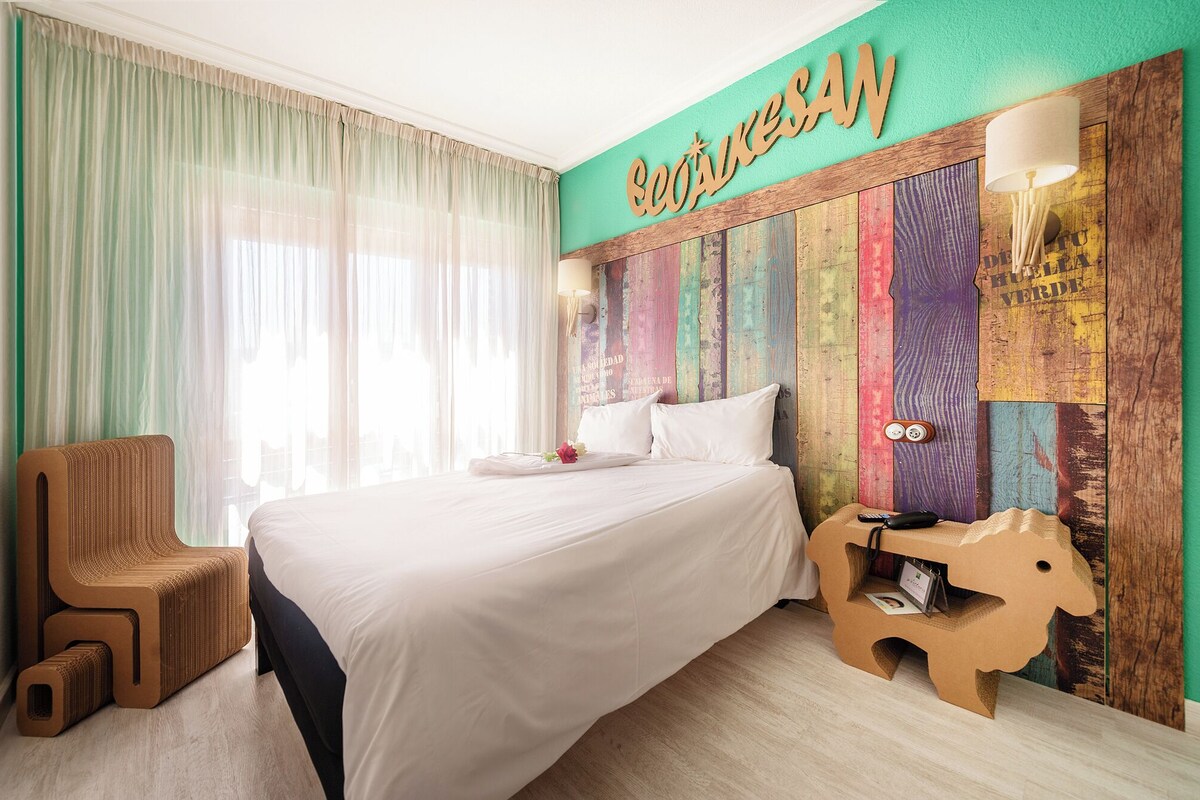 A hotel room with a bed, a wooden headboard with colorful panels, a wooden side table, and a chair.