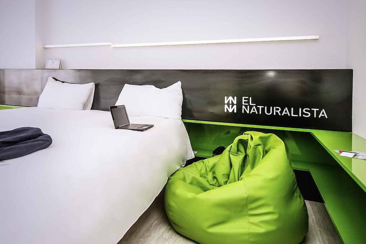 A modern hotel room with a bed, a green bean bag chair, and a headboard with the text "EL NATURALISTA".