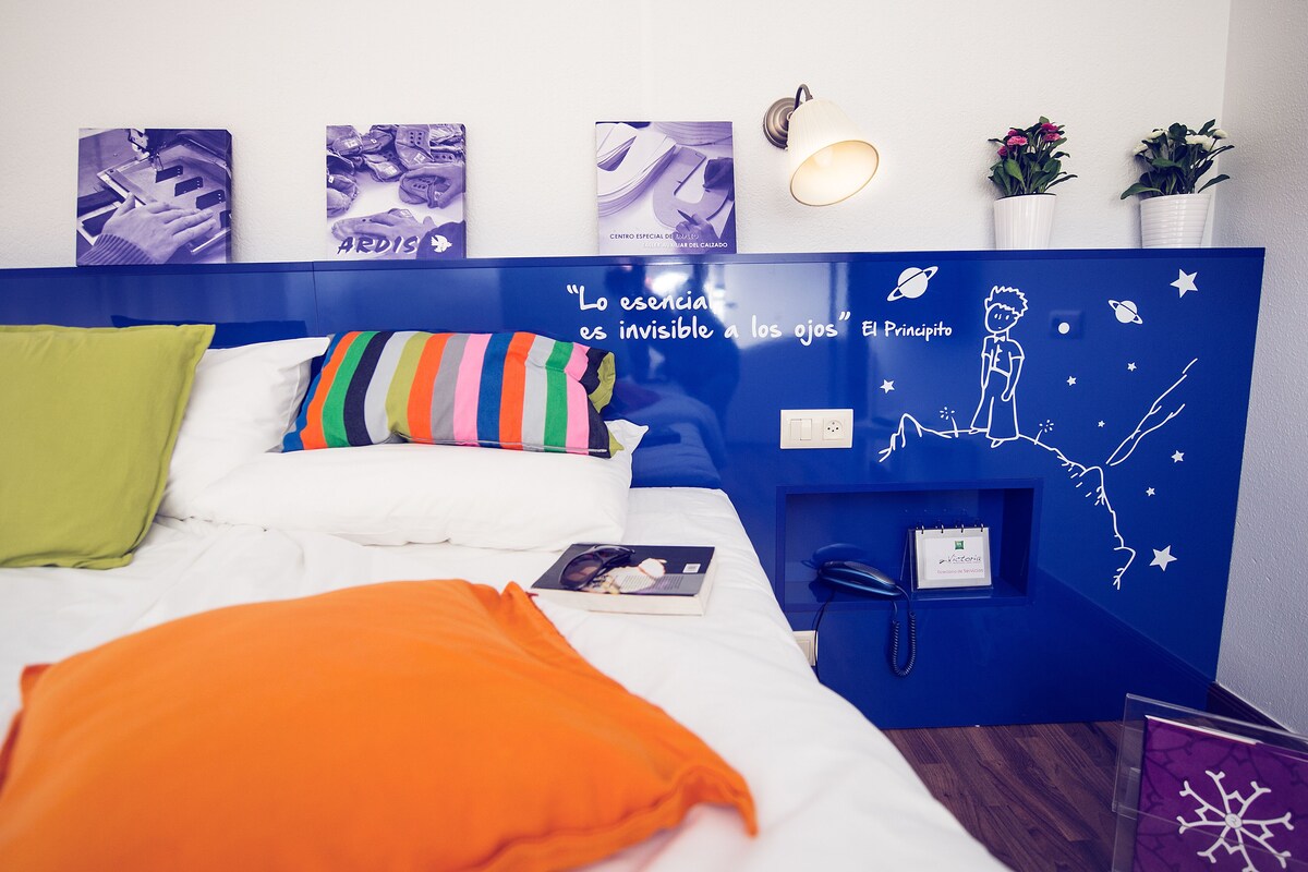 Un dormitorio con una cama, almohadas, una mesita de noche, una pared decorada con obras de arte, una lámpara de pared y un estante con un teléfono y una tarjeta.