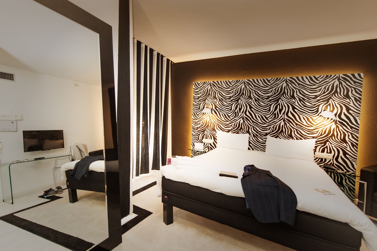 A modern hotel room with a zebra-patterned headboard, a flat-screen TV mounted on the wall, a bedside table with a lamp, and a neatly made bed.