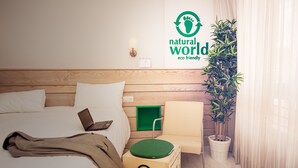 Habitación de hotel con cabecera de madera, cama con sábanas blancas, una computadora portátil sobre la cama, una silla verde, una planta en maceta y una pared con un letrero que dice 'mundo natural, eco-friendly'.