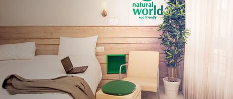 Habitación de hotel con cabecera de madera, cama con sábanas blancas, una computadora portátil sobre la cama, una silla verde, una planta en maceta y una pared con un letrero que dice 'mundo natural, eco-friendly'.