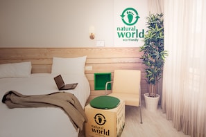 Habitación de hotel con cabecera de madera, cama con sábanas blancas, una computadora portátil sobre la cama, una silla verde, una planta en maceta y una pared con un letrero que dice 'mundo natural, eco-friendly'.