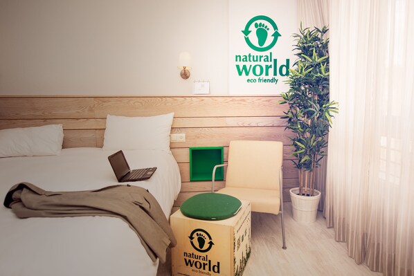 A hotel room with a wooden headboard, a bed with white linens, a laptop on the bed, a green chair, a potted plant, and a wall with a 'natural world eco friendly' sign. - ibis Styles La Rioja Arnedo (Arnedo)