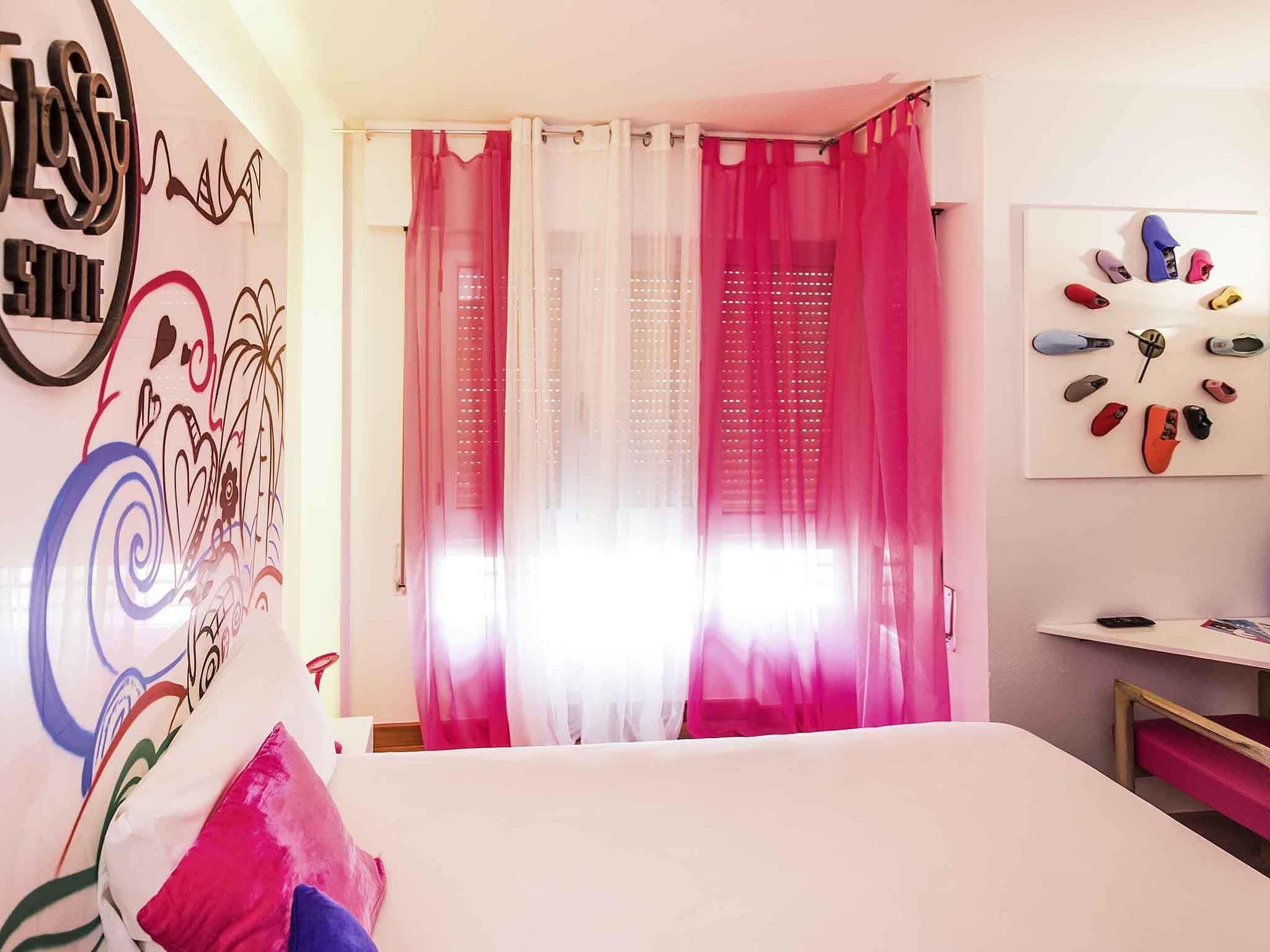 Un dormitorio con cama, cortinas rosadas, una pared decorada con corazones y remolinos, y un zapatero con varios pares de zapatillas.