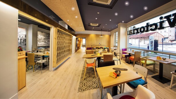 Una cafetería moderna con muebles de madera, una alfombra estampada y vistas a la calle a través de amplios ventanales.