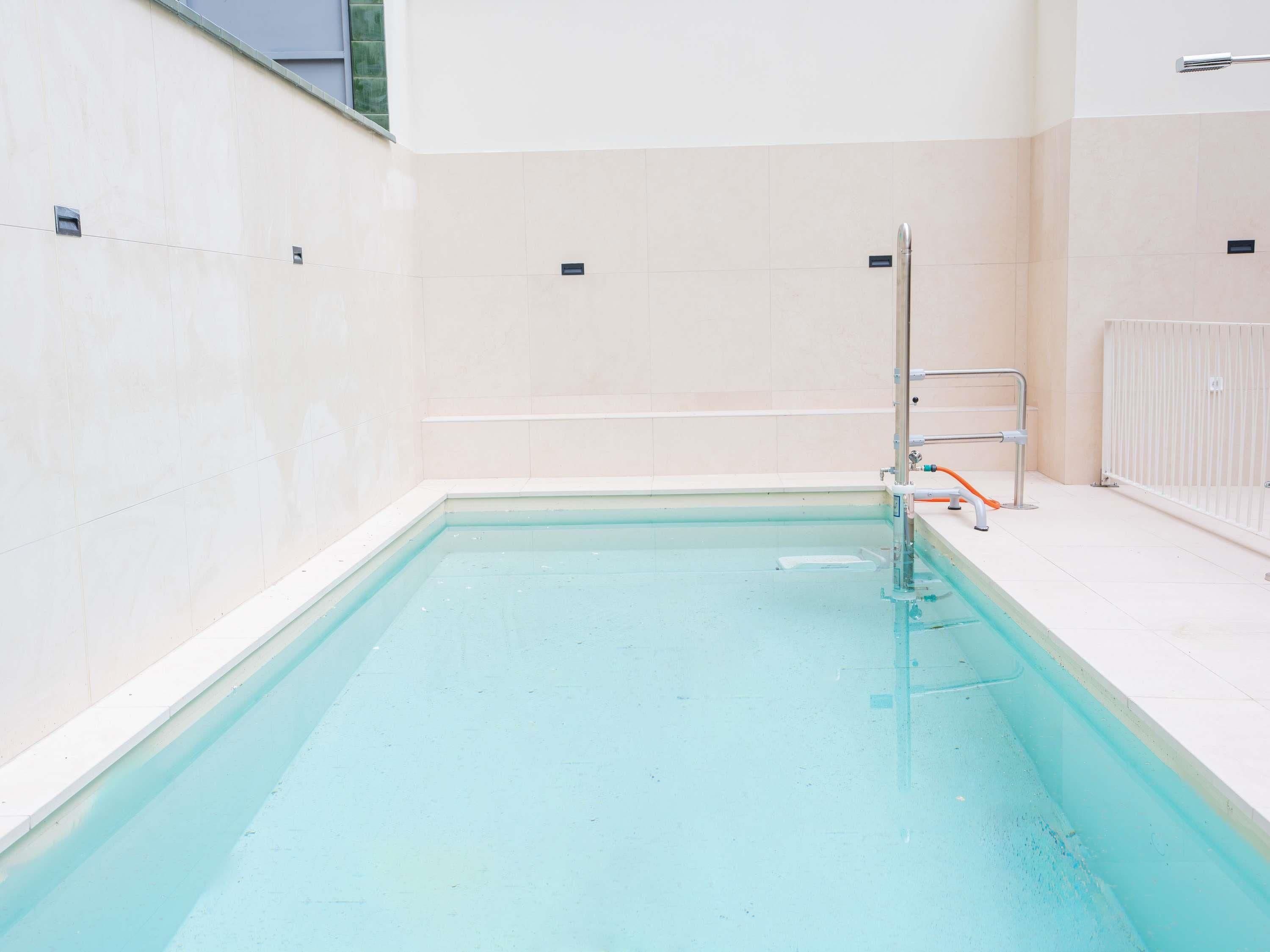 Una piscina con agua cristalina azul, rodeada de paredes de baldosas blancas y una escalera metálica.