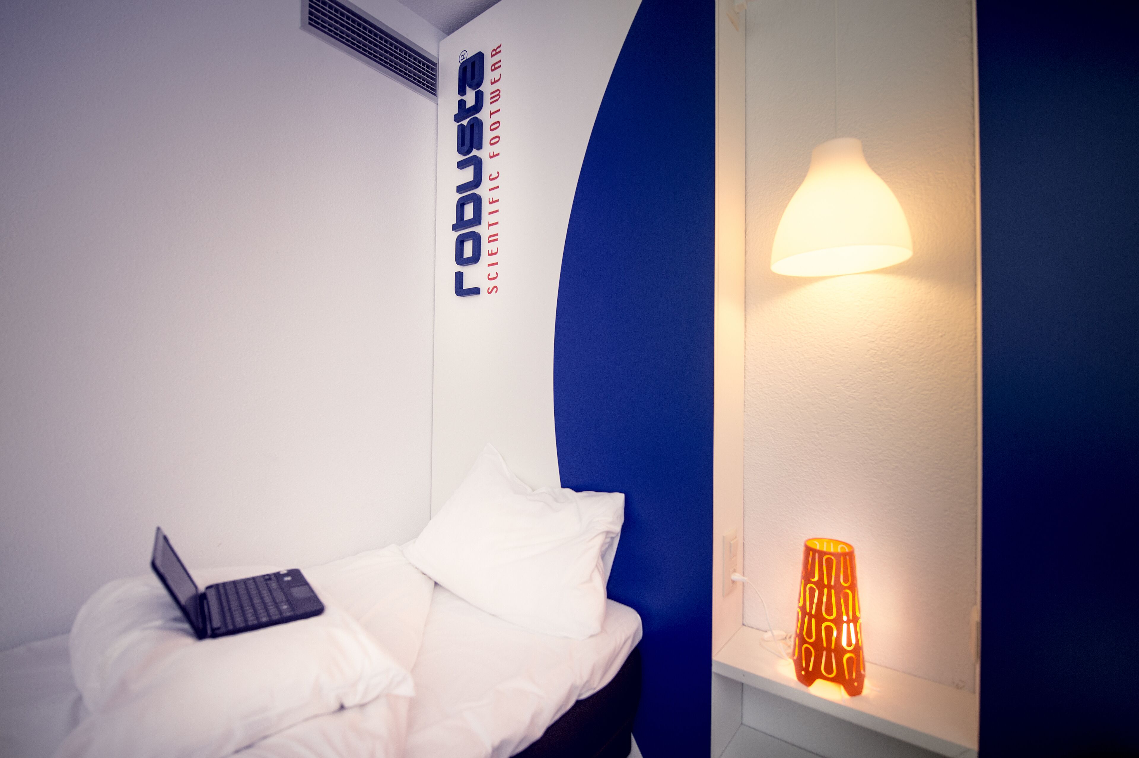 A hotel room with a bed, a laptop, a wall-mounted air vent, and a wall-mounted light fixture.