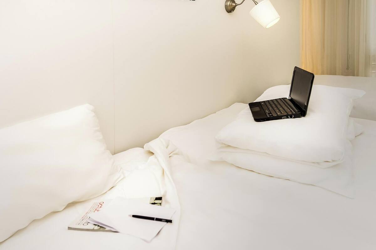 A hotel room with a bed, a laptop on the bed, and a lamp on the wall.