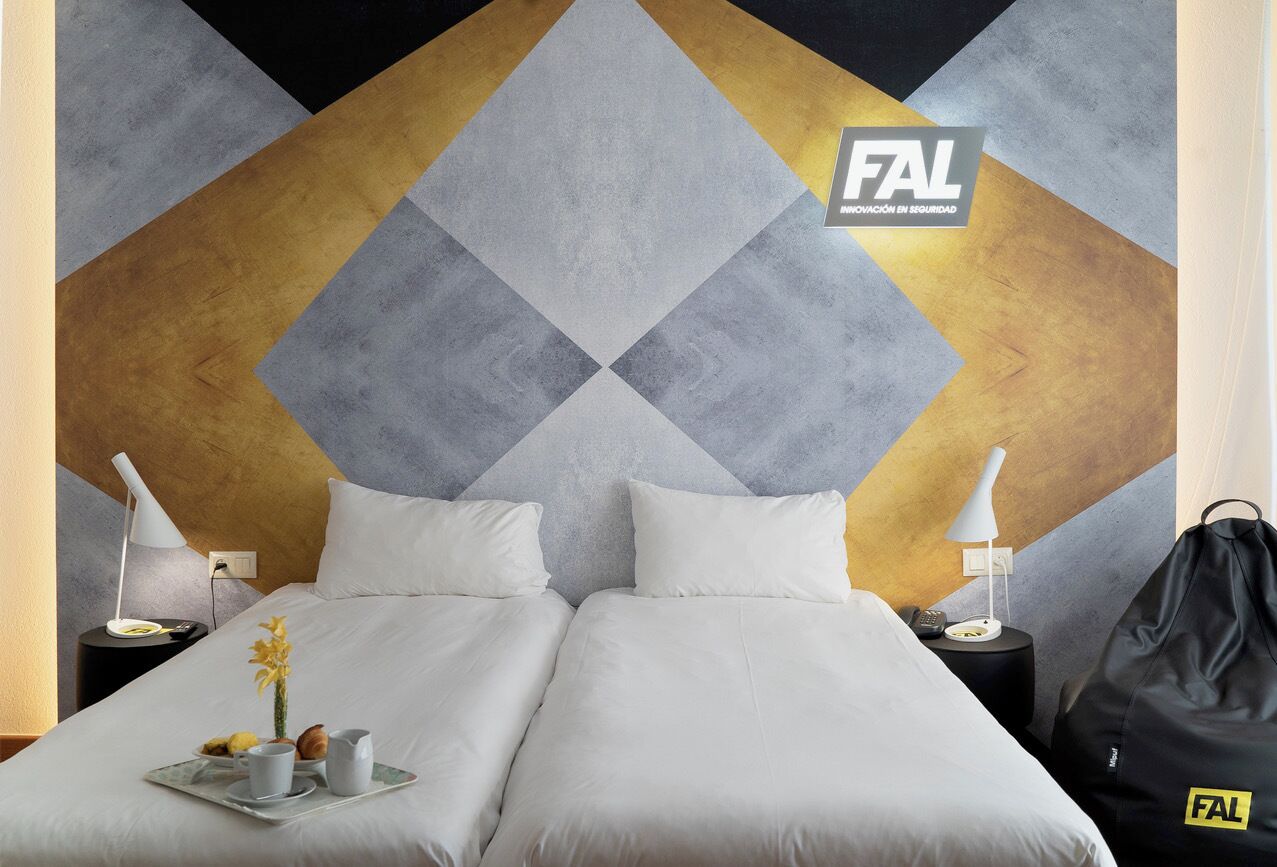 Habitación de hotel con dos camas, un diseño geométrico en la pared, una bandeja con alimentos para el desayuno y un logotipo en la pared.