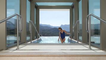 Una mujer con traje de baño azul al borde de una piscina infinita de vidrio con vista a la montaña.