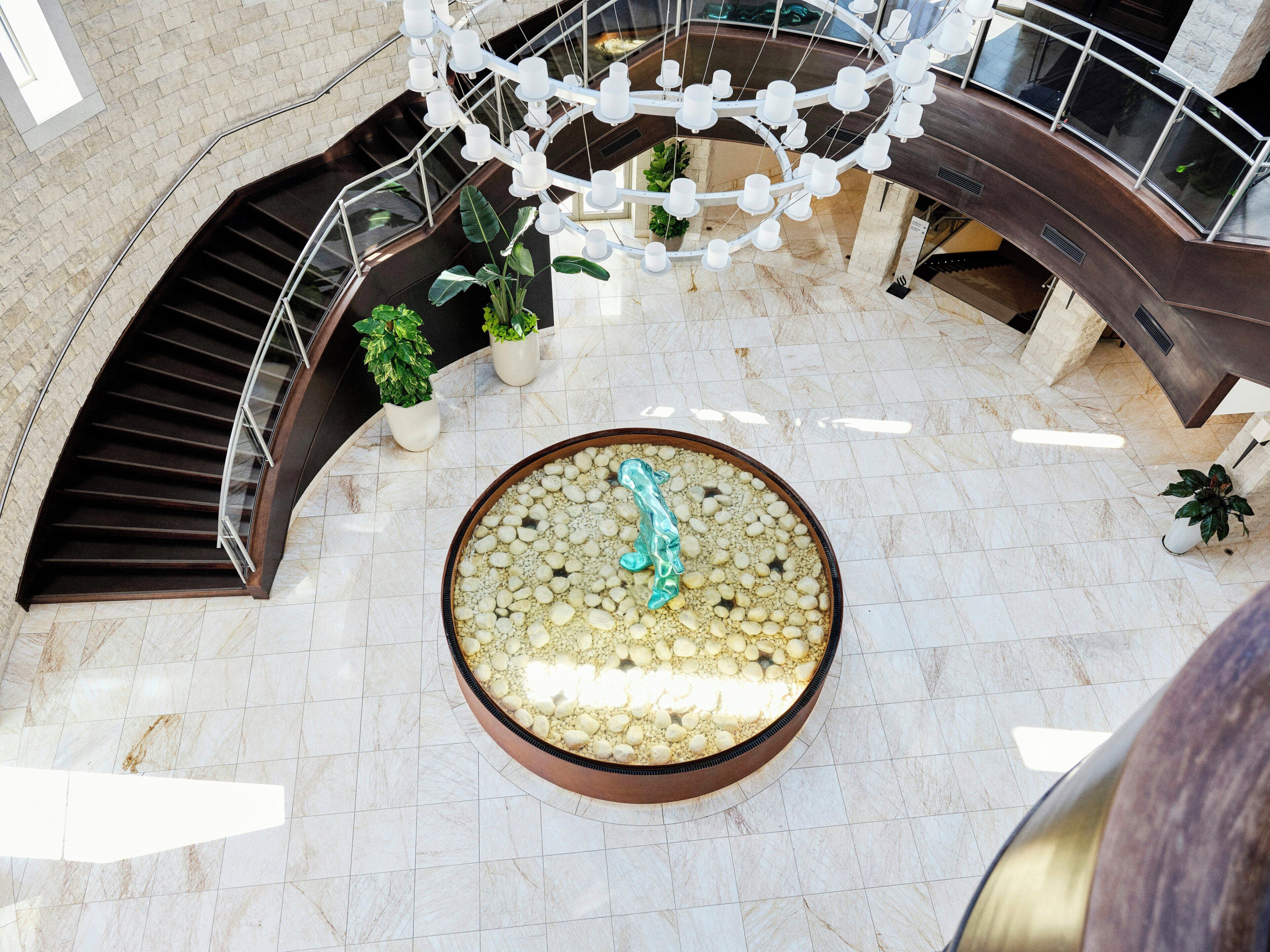 A circular water feature with pebbles and a central sculpture, surrounded by a marble floor and potted plants.