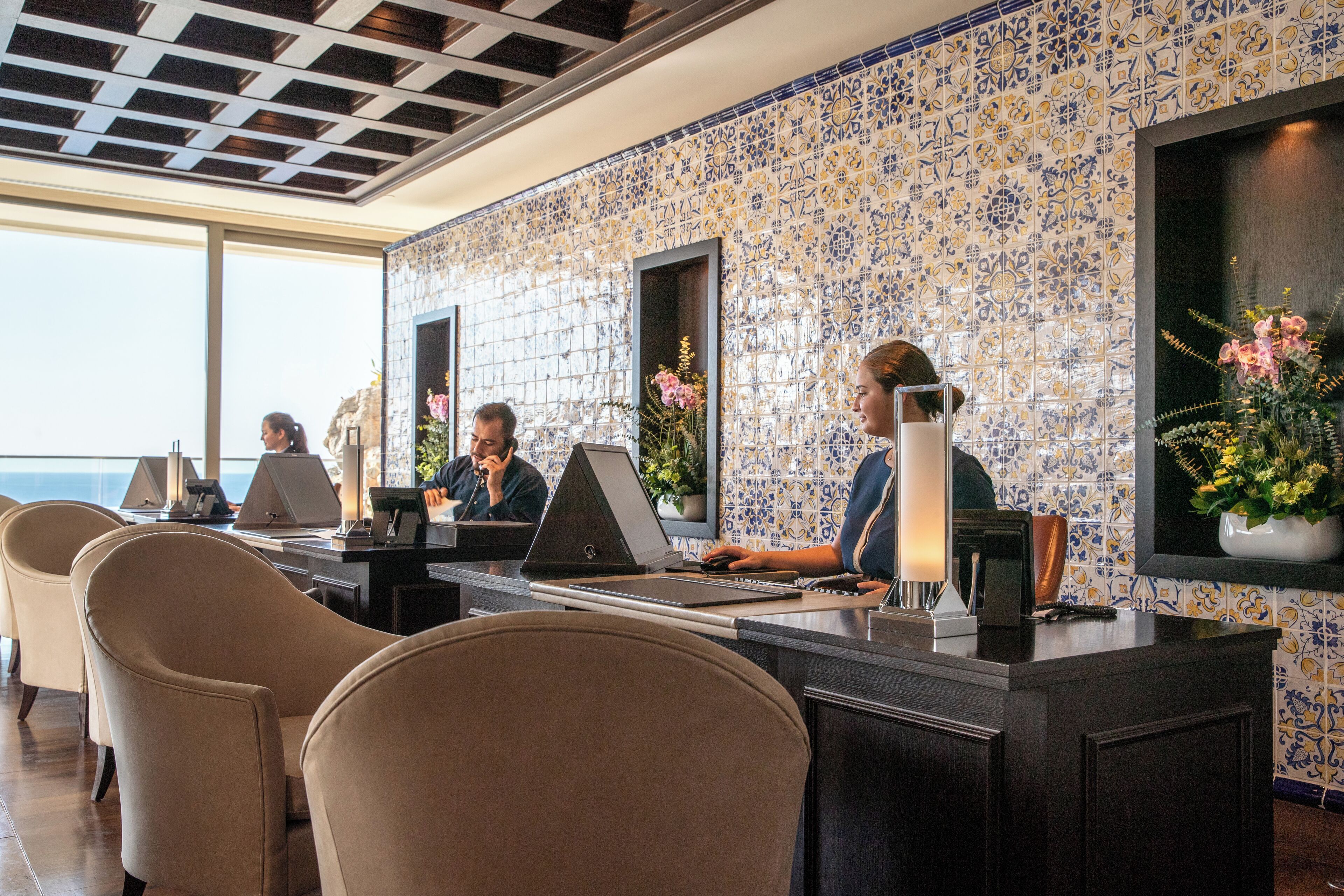 Un mostrador de recepción de hotel con varias estaciones de trabajo, un amplio ventanal con vistas al mar y una pared de azulejos con diseño floral.