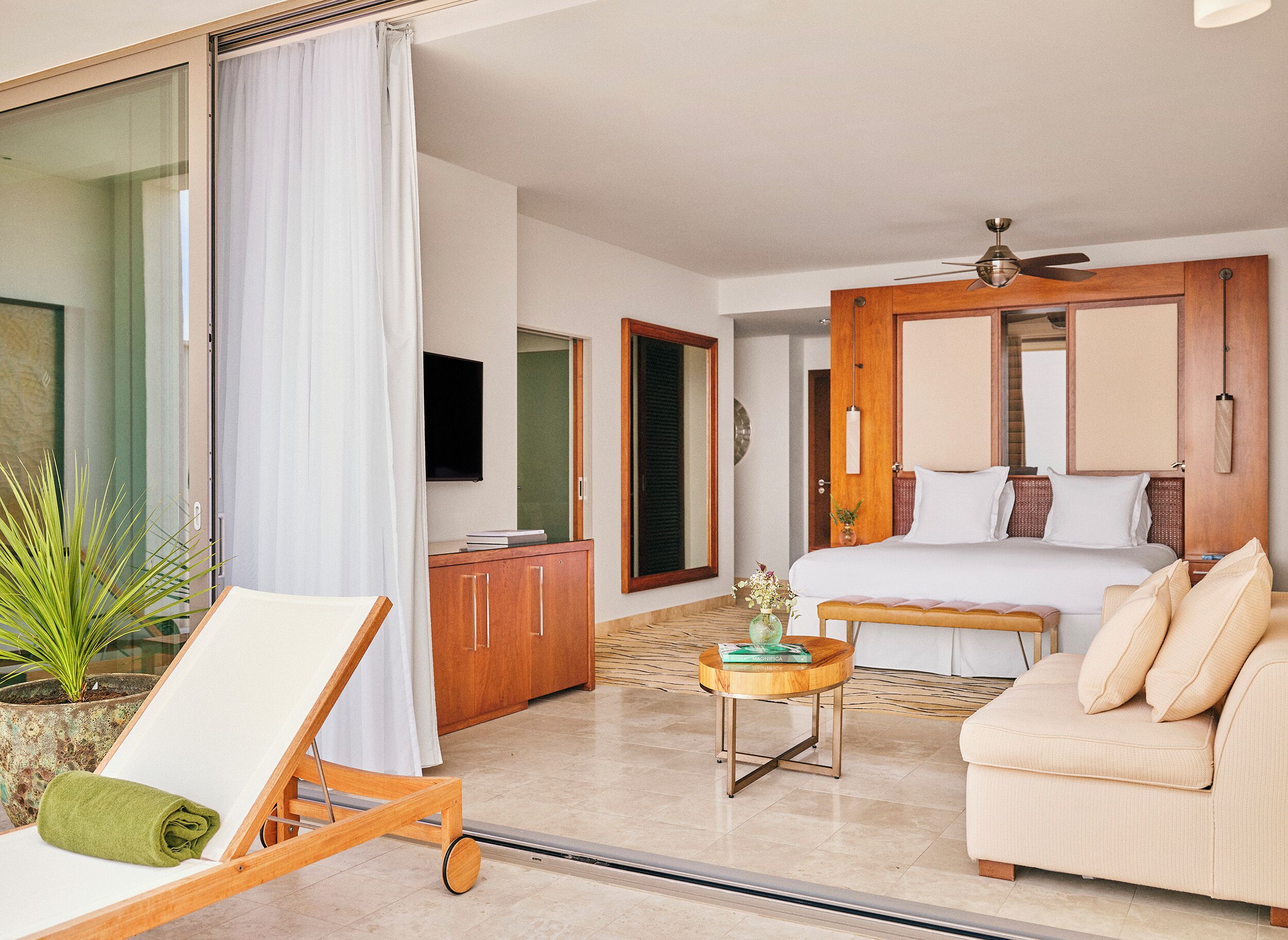 A hotel room with a bed, a sofa, a chair, a small table, a TV, a ceiling fan, and a potted plant.