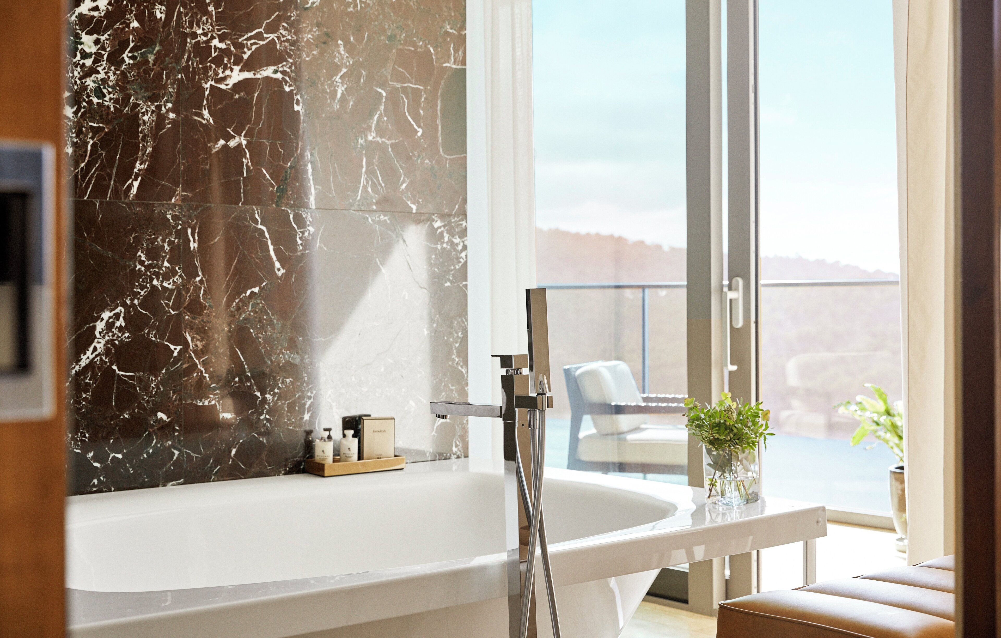 A modern bathroom with a marble wall, a bathtub, and a glass-enclosed balcony with a view.
