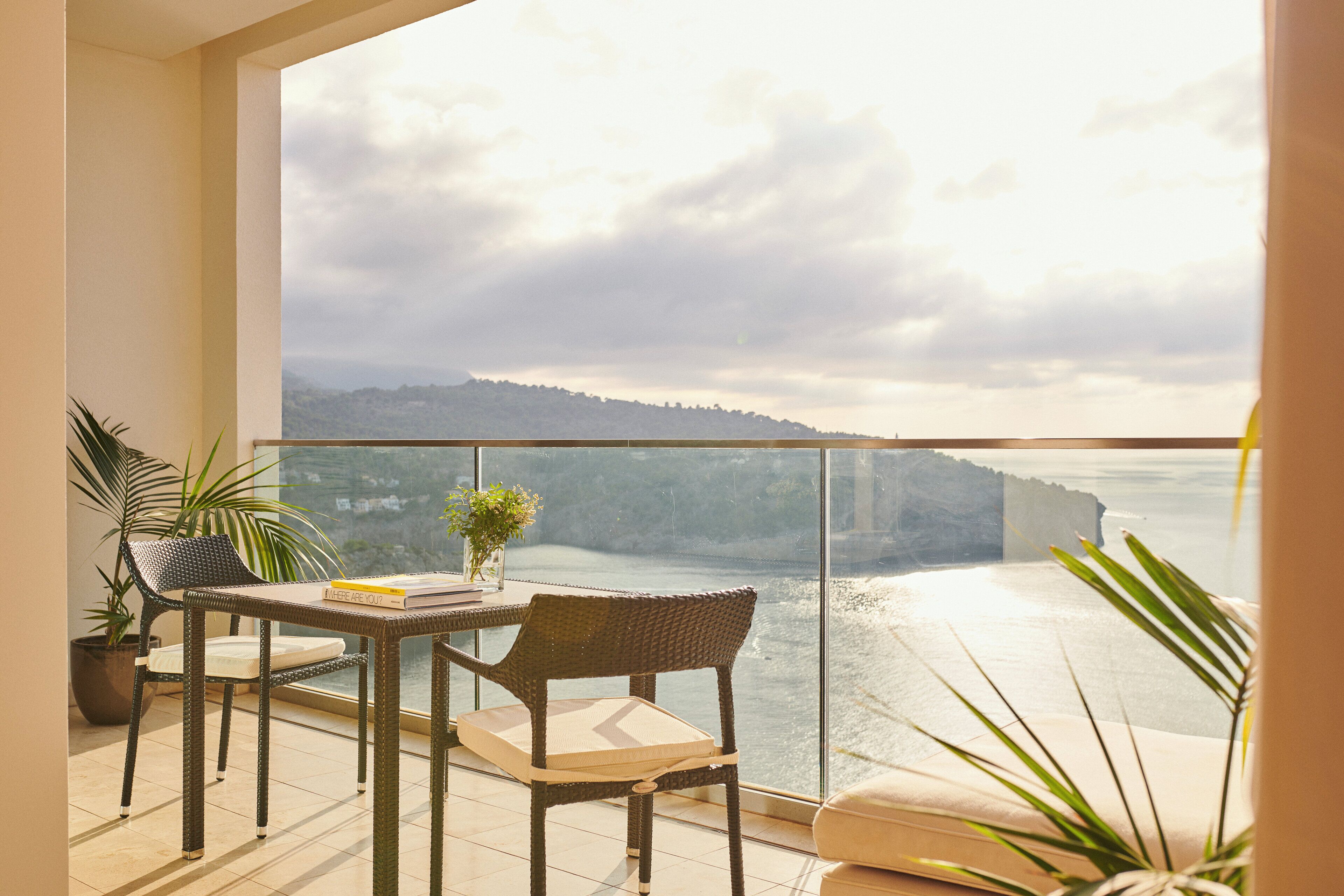 A balcony with a view of a coastal landscape, a table with chairs, and potted plants.