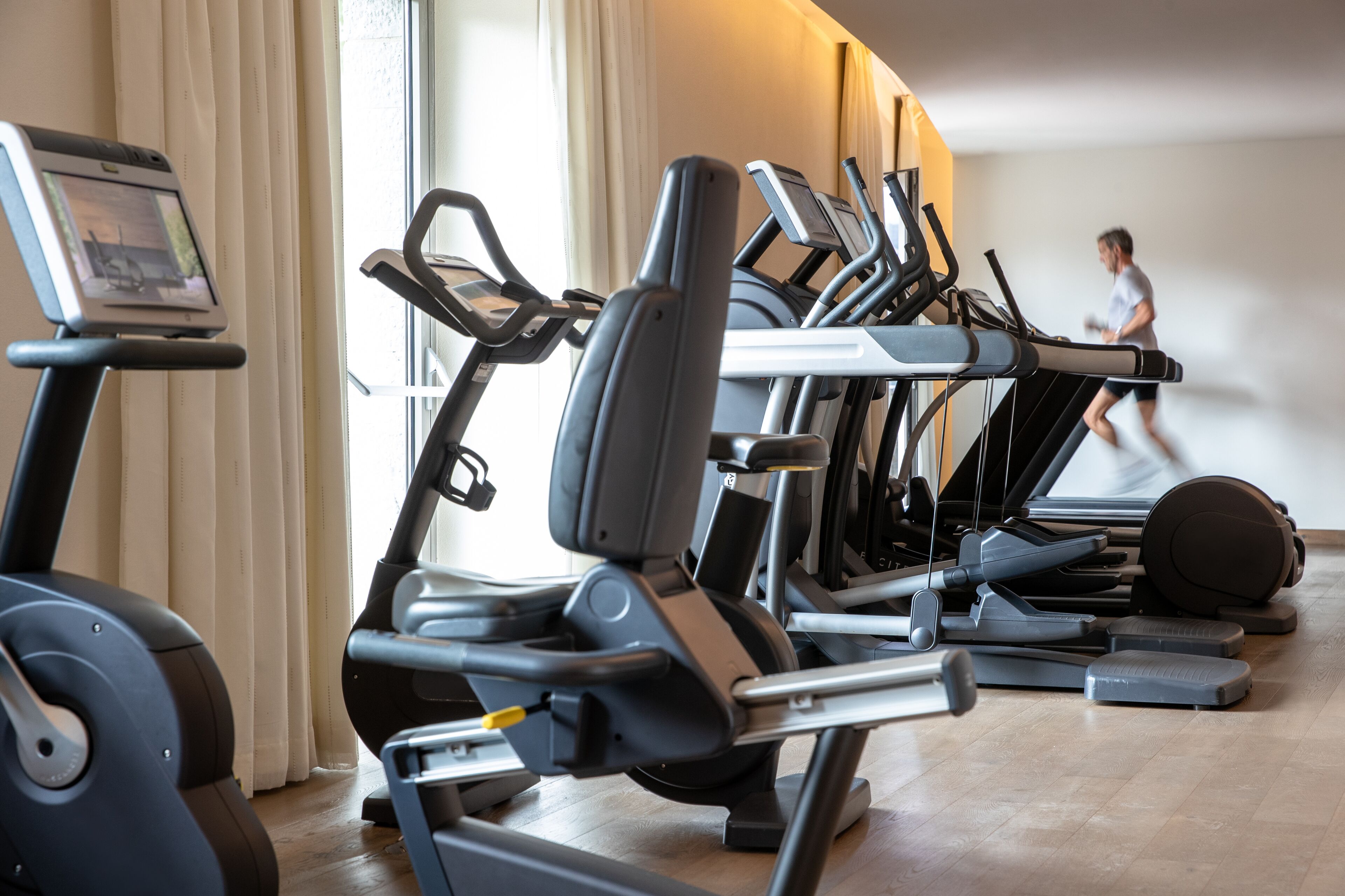 A row of stationary bikes and treadmills in a gym with a person running in the background.