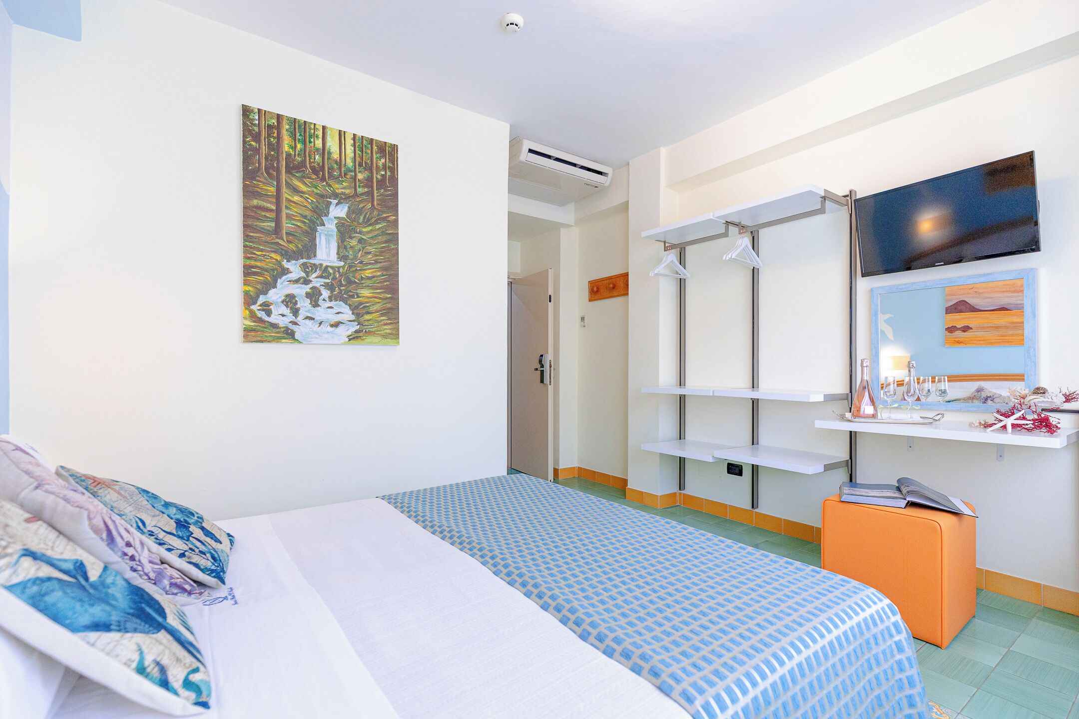 A hotel room with a bed, a painting of a waterfall, a TV, a shelf with toiletries, and a small orange ottoman.