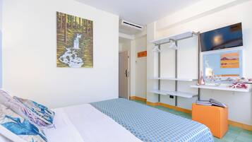 A hotel room with a bed, a painting of a waterfall, a TV, a shelf with toiletries, and a small orange ottoman.