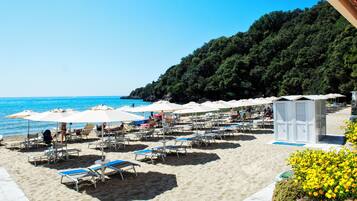 A beach with rows of sun loungers and umbrellas, a small kiosk, and a forested hill in the background.
